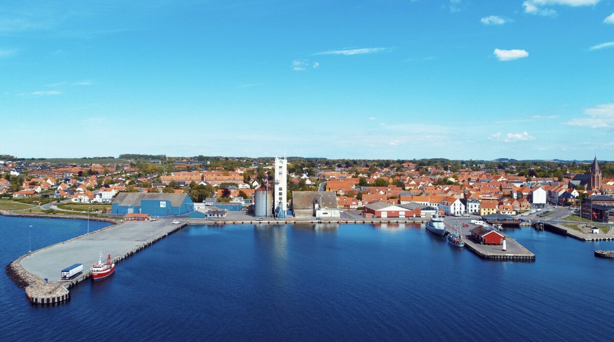 Assens kyst og havn set ovenfra. Tre både ligger i havnen, bygninger på landjorden ses i baggrunden.