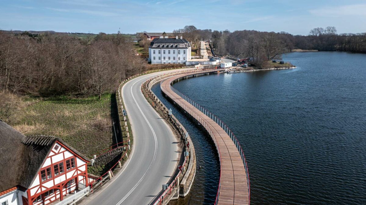 Cykelstibro med træ på overfladen slanger sig over søen og langs vejen ved Hvidkilde Gods. Der er en stribe vand mellem vej og bro. Det hvide gods ligger for enden af broen