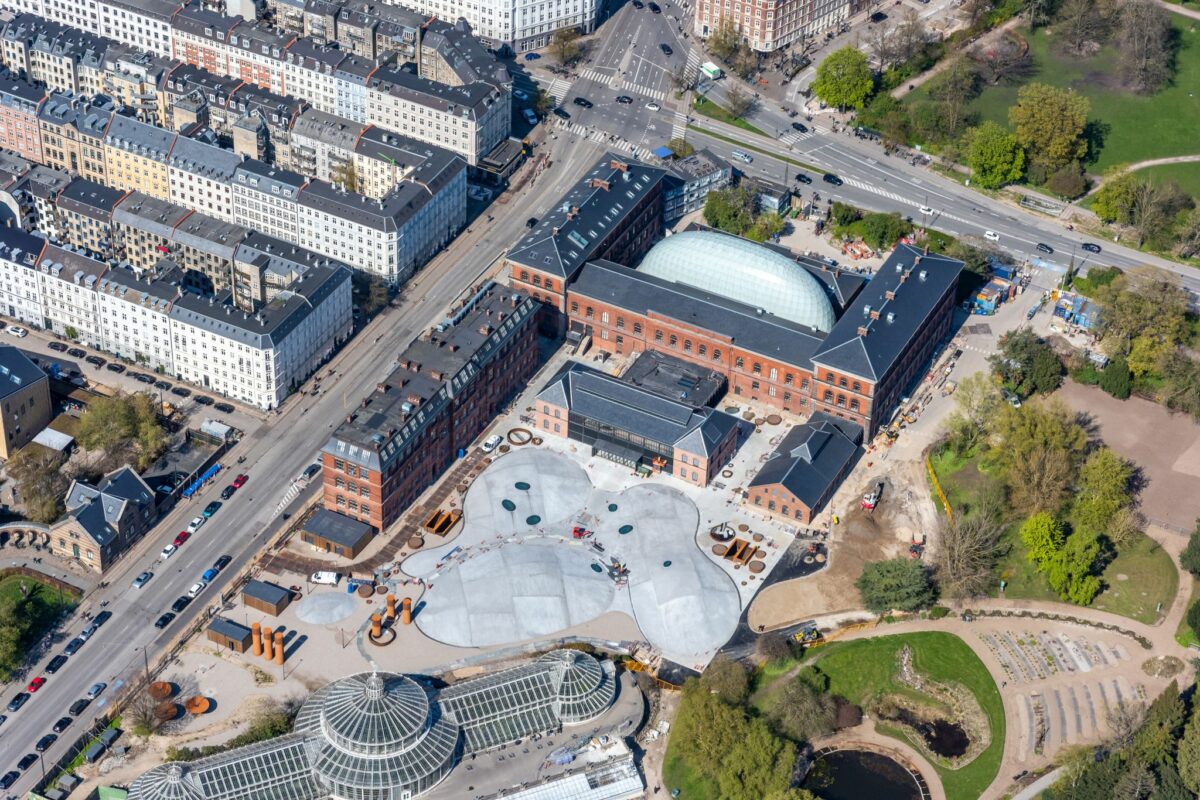 Luftfoto viser de gamle røde bygninger med sort tag - bl.a. omkranser tre længer den nye kuppel i metal og glas. Foran hovedindgangen er små, grå bakker. til højre Botanisk Haves grønne planter og træer. I forgrunden det store drivhus.