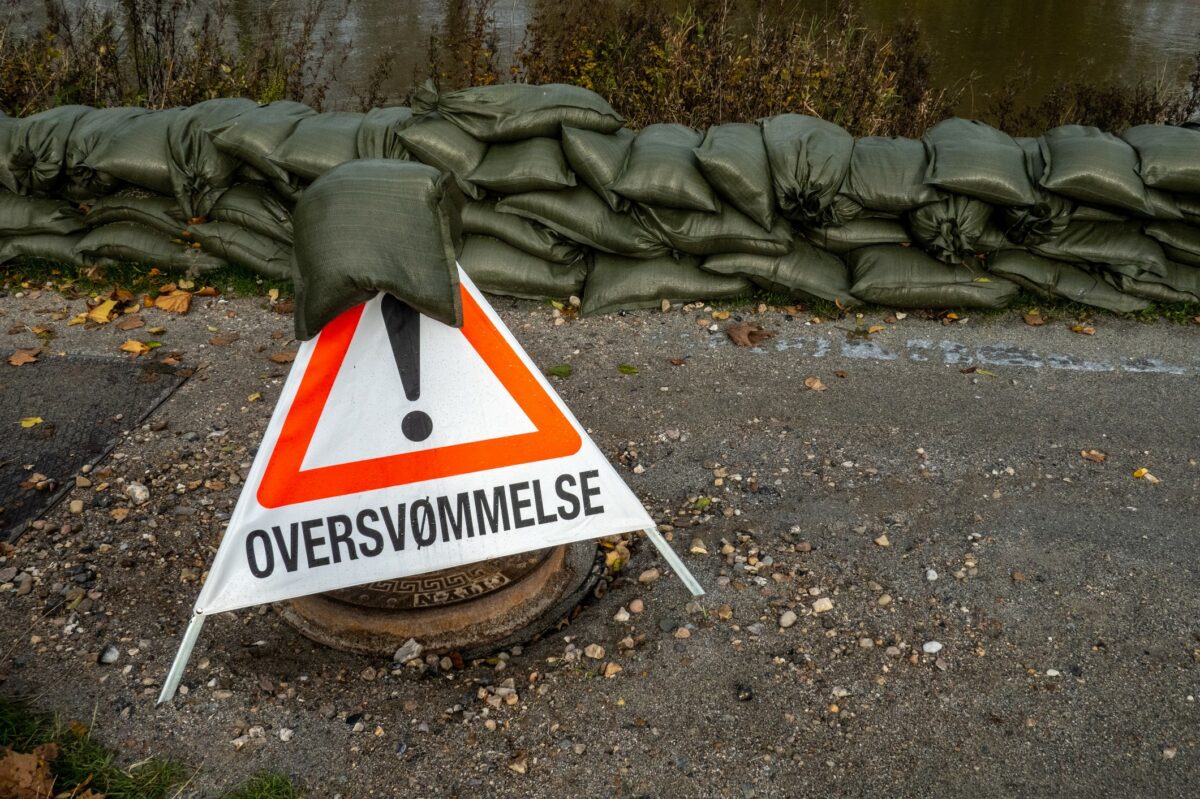 Et trekantet skilt med Oversvømmelse står på en vej foran en lav mur af grønne sandsække