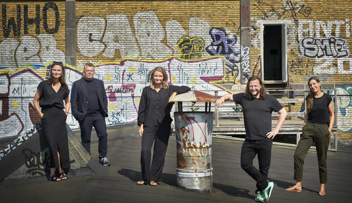 Fem mennesker, klædt hovedsageligt i sort, fotograferes foran væg malet med graffiti.