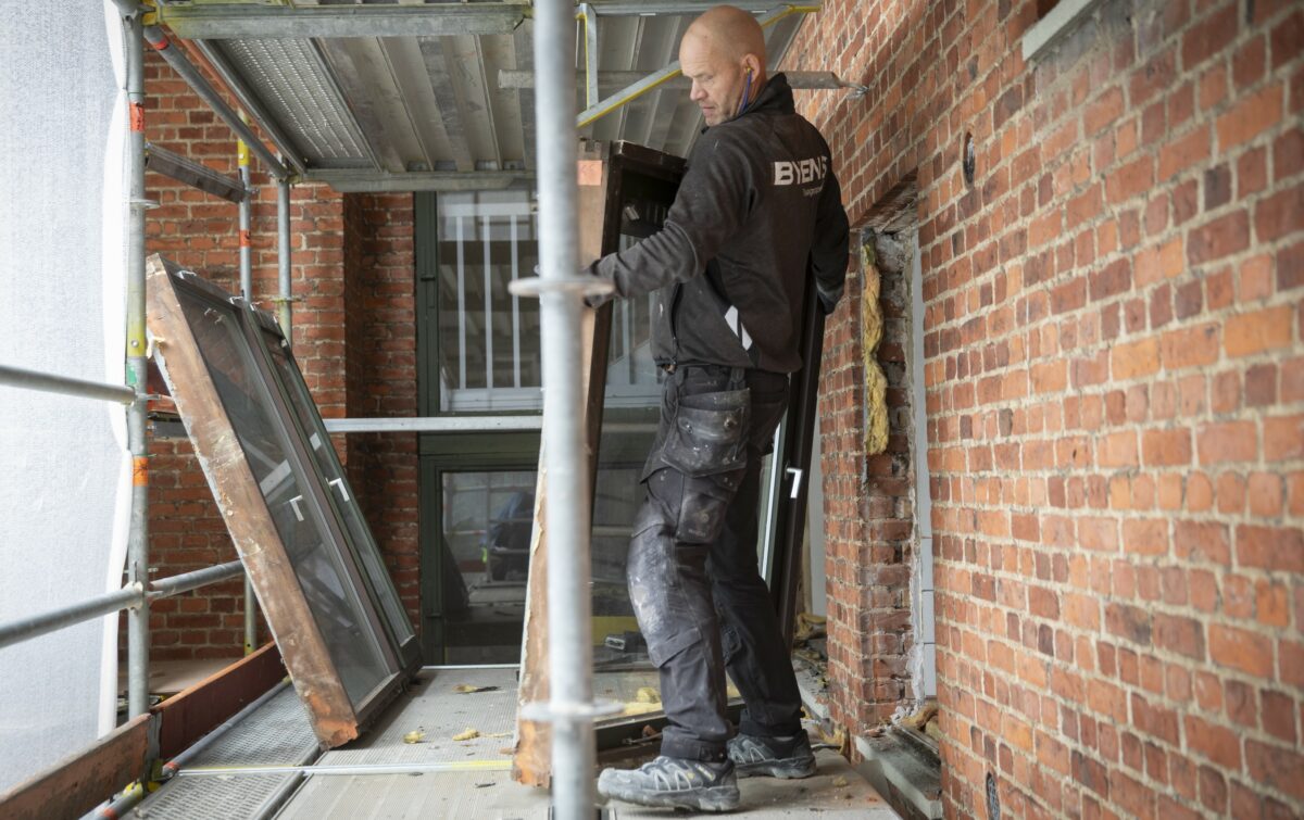 En tømrer i sort arbejdstøj, sorte og støvede sikkerhedssko og med blå høreværn i ørerne bugserer et gammelt vindue ud af et vindueshul. Han står på et stillads. Til venstre en hvid presenning. Til højre muren i røde mursten.