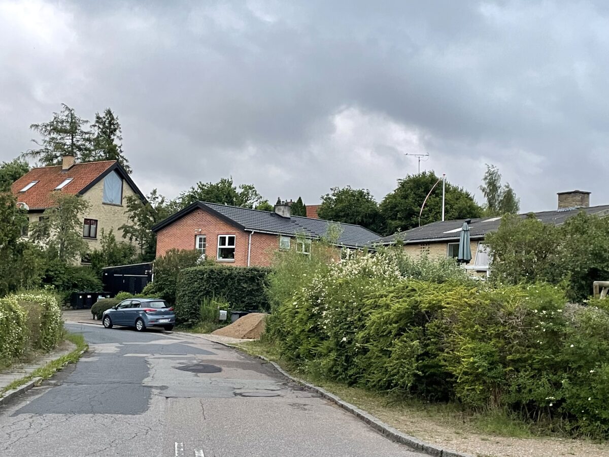 Foto ned ad en vej, hvor der ligger flere boliger i 1-3 plan i hvid, lyserød og hvid. Der holder biler parkeret i bunden af vejen