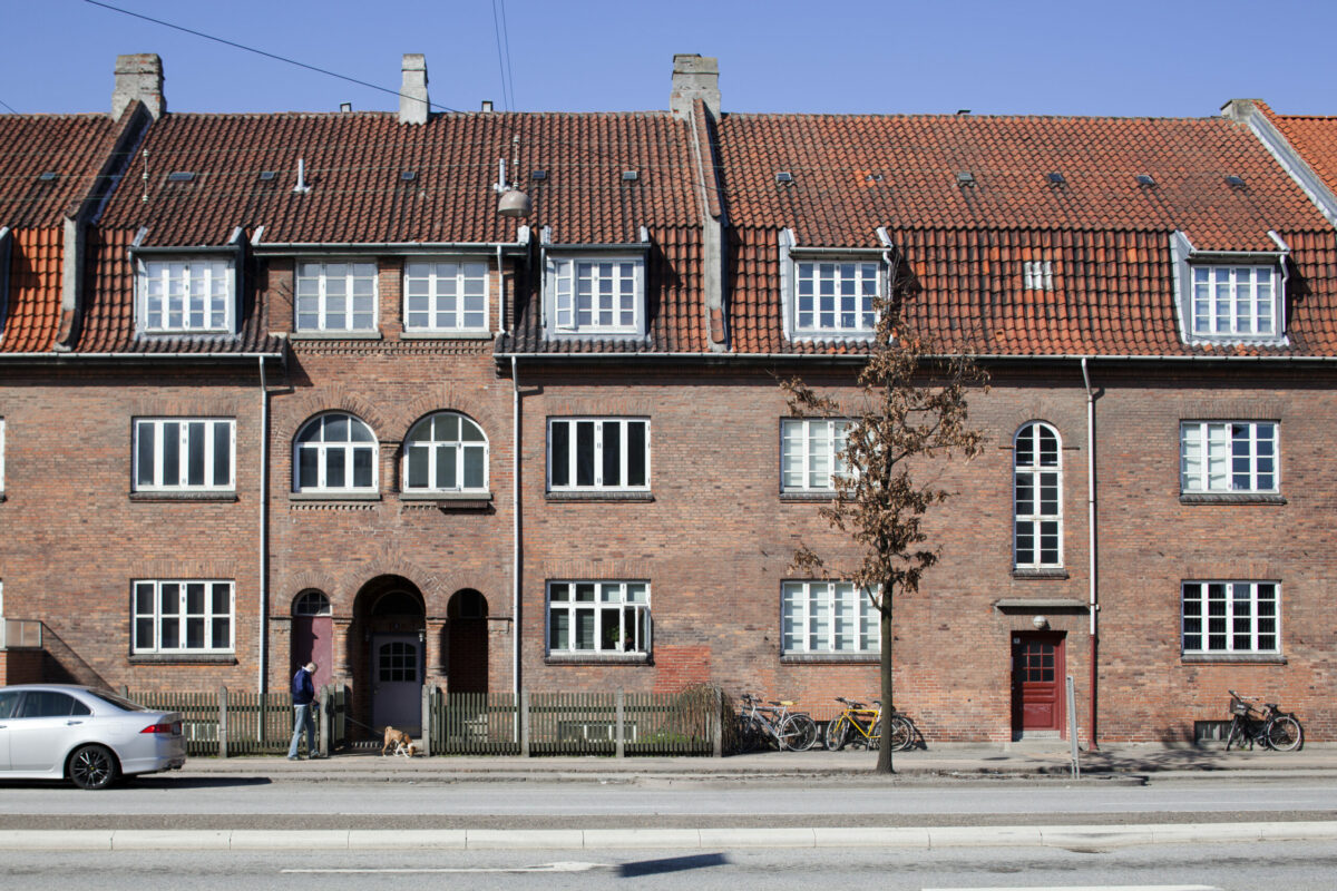 Foto af en ældre rød bebyggelse i to etager og med boliger i loftetagen også. Foran bygningen på gaden går en mand. En hvid bil er parkeret. Opgangsdøren er rød.