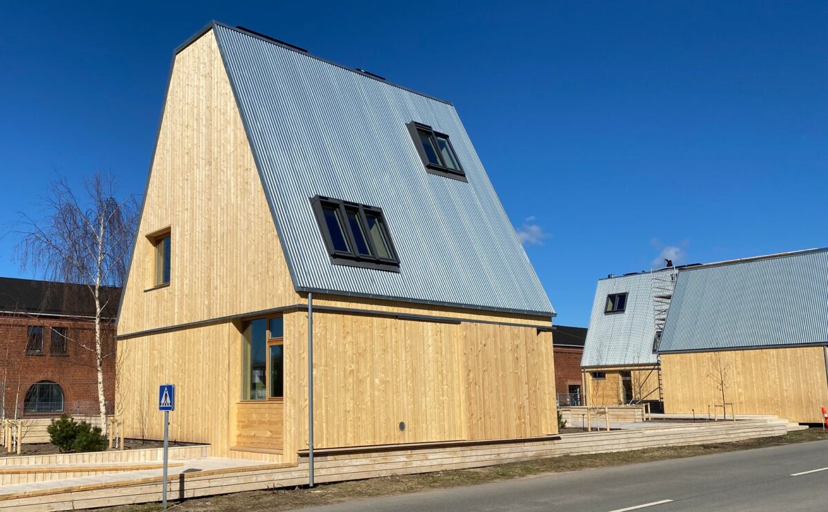 Træhus tæt på - høj rejsning på taget, der er gråt. I baggrunden ses flere huse.