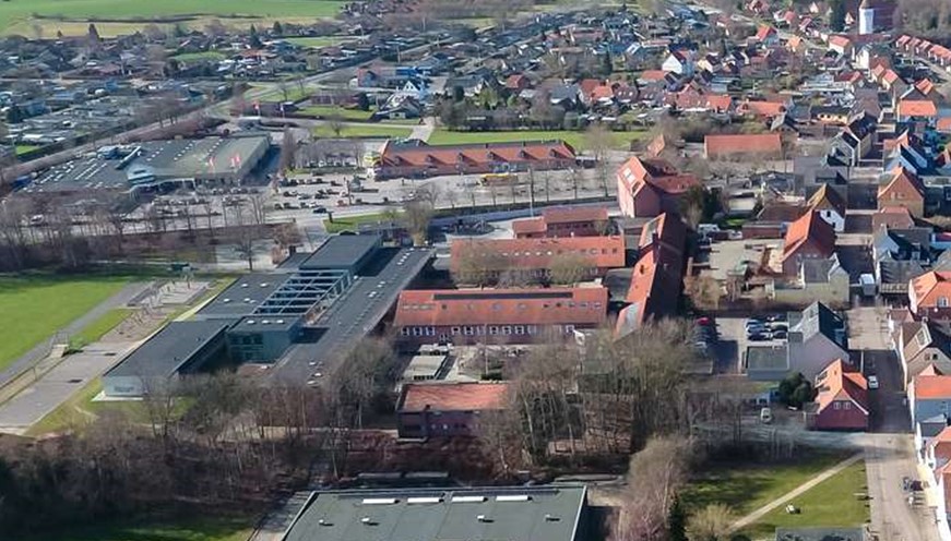 Luftfoto over Nordborg med lange, røde skolefløje i midten. I forgrunden en firkantet, sort kasse af en bygning