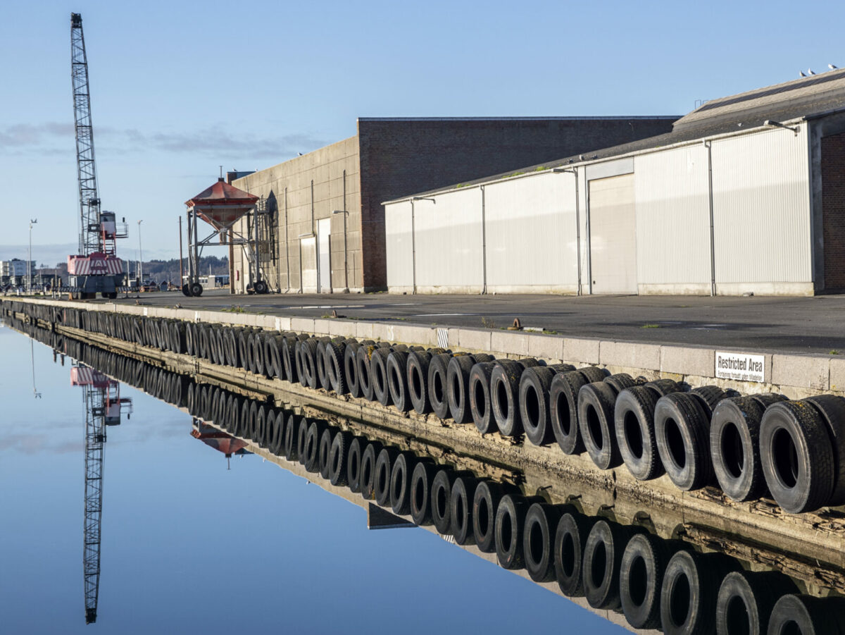 Foto af grå bygninger lige vej kajkanten. Der er bildæk på kajsiden ud mod havnen. I baggrunden en kran.