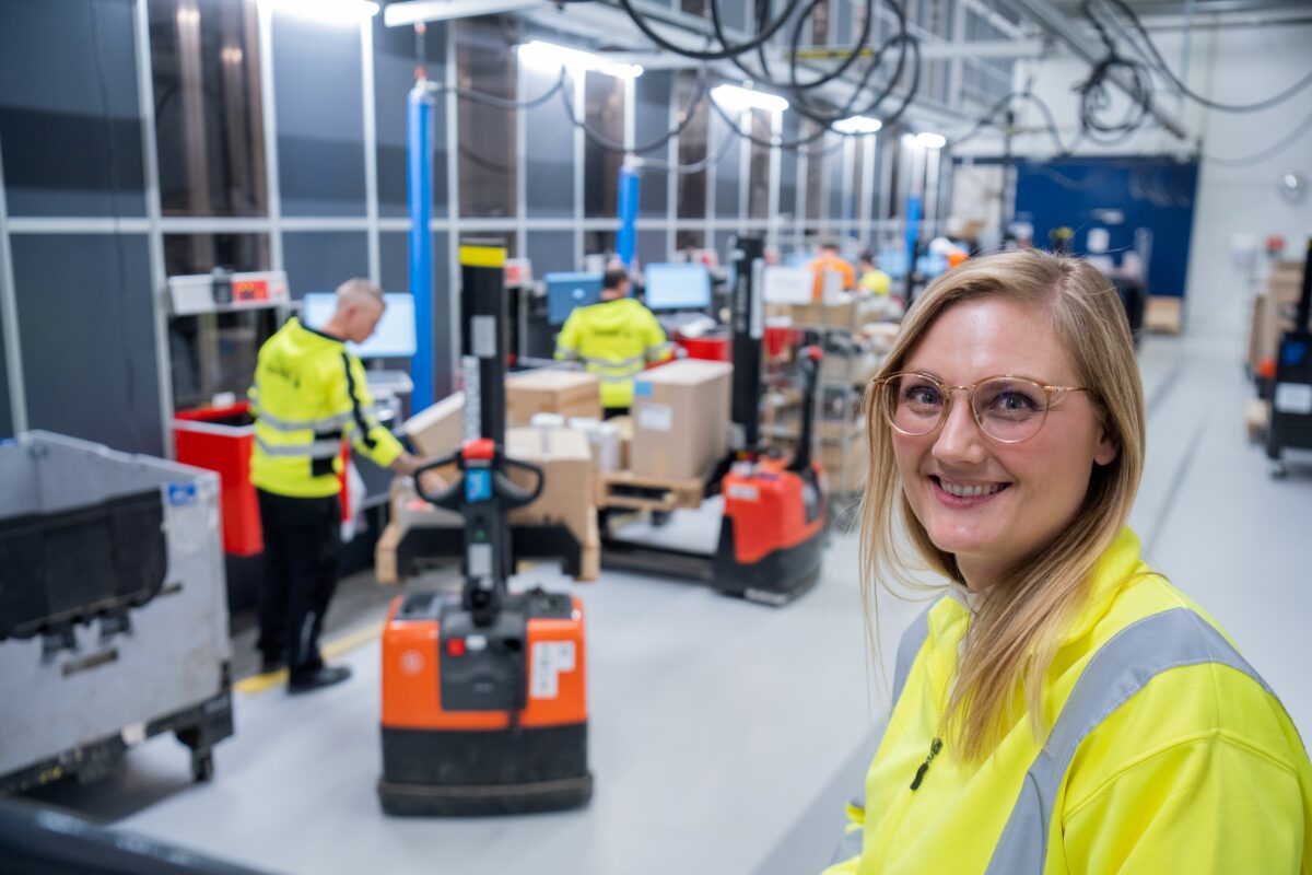 Kvinde i refleksjakke smiler til kameraet, i baggrunden står flere mennesker i refleksjakker med palleløftere.