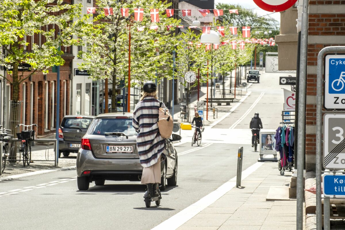 Biler, cykler og el-løbehjul færdes på gade med butikker i Esbjerg.