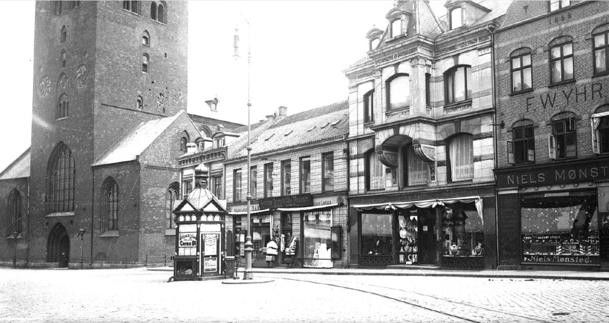 sort/hvid foto af den nederste del af Aarhus Domkirke - på torvet ses en aviskiosk og flere butikker