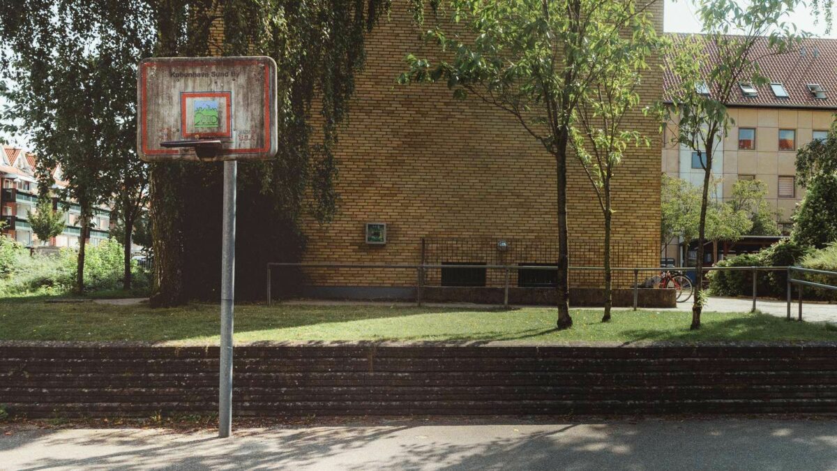 Foto af gavlen på en bygning bygget i gule mursten. Foran bygningen står lidt træer. Tættest på kameraet en meget beskidt basketkurv på en metalstang