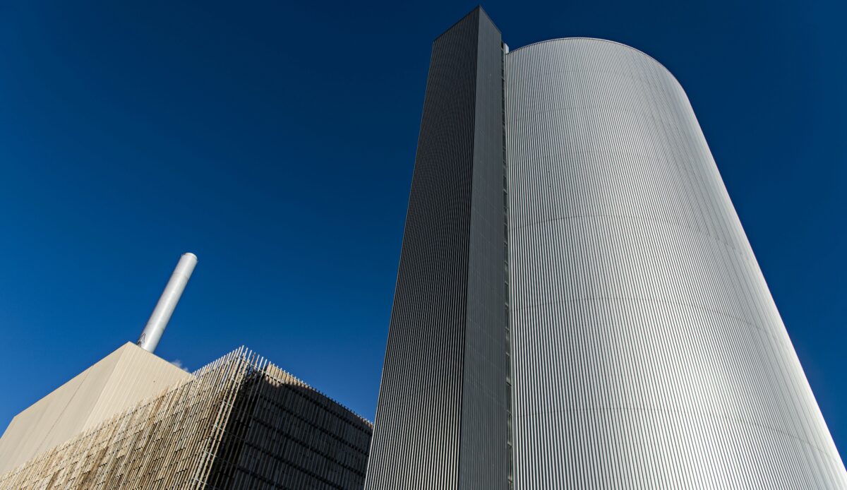 Kig op ad en stor silo på Amagerværket. Til venstre ses en højere skorsten ovenpå en betonbygning