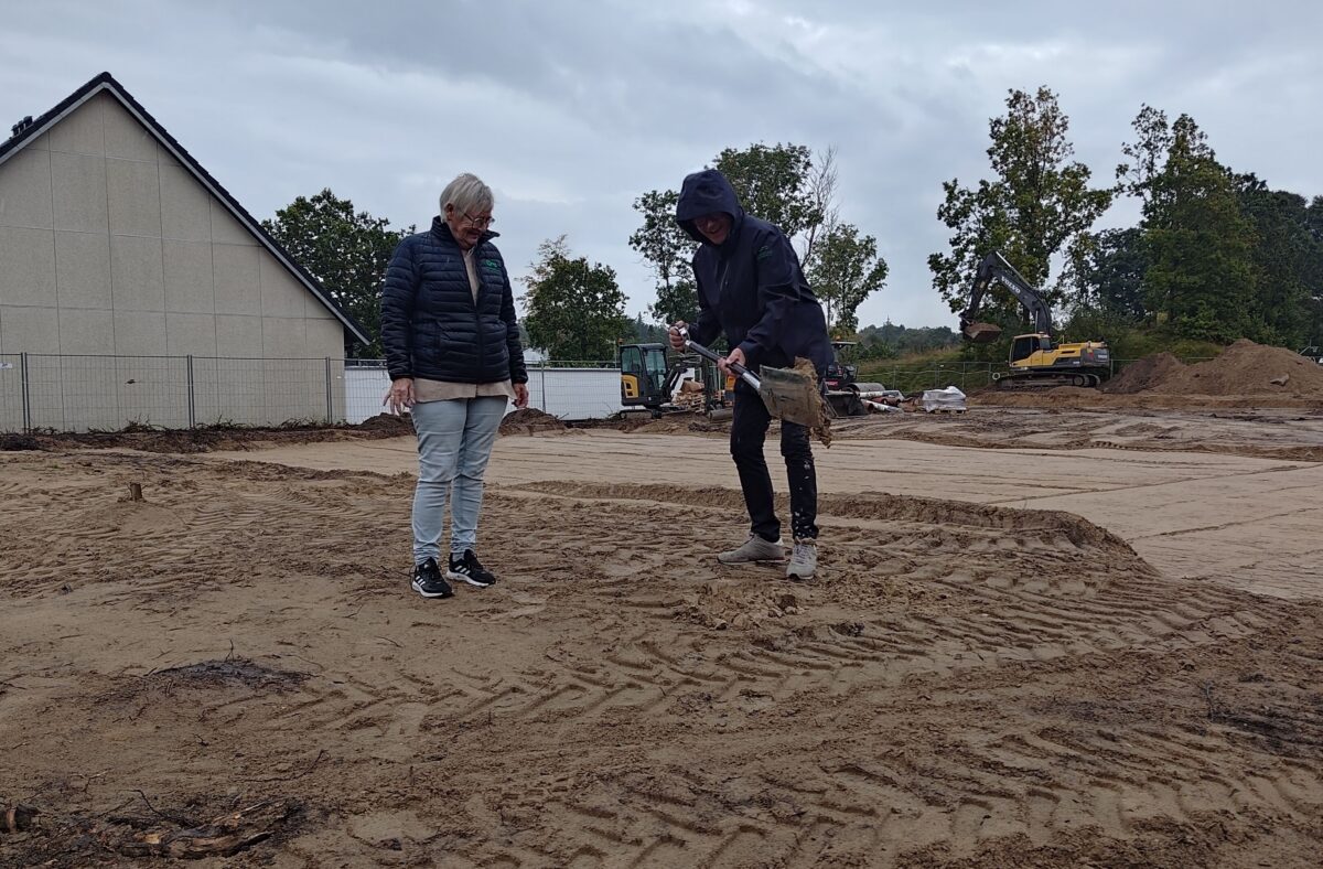 To mennesker - den ene med hætten oppe og en spade i jorden - tager første spadestik på ni nye rækkehuse. Bag ses gavlen på en hvid bygning