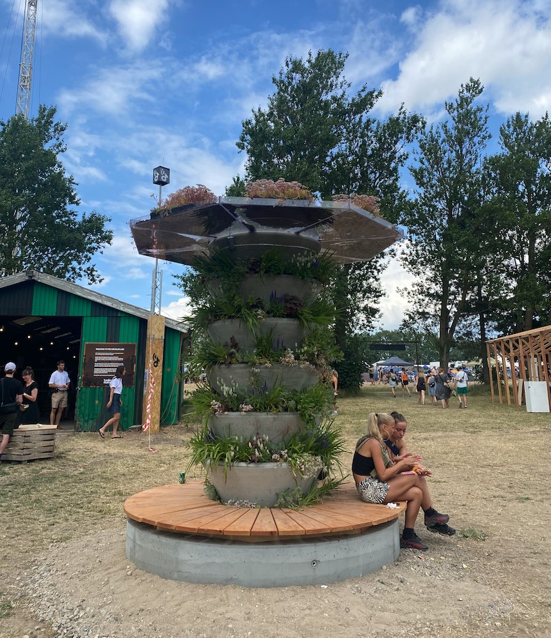 To piger med bare ben sidder på bænken i træ, der går hele vejen rundt for foden af Urban Tree. I baggrunden til venstre ses et grønt hus. Til højre går en flok mennesker hen mod festivalpladsen