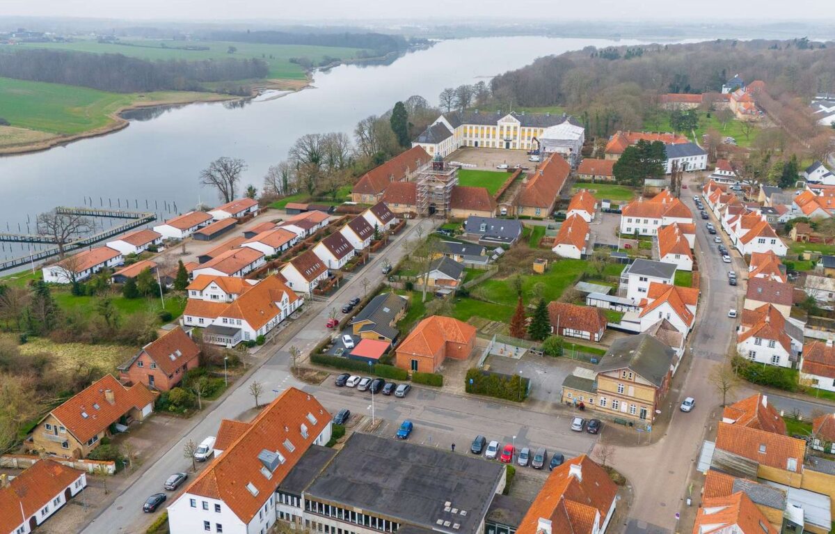 luftfoto af Augustenborg med røde tage og hvide huse. Der løber vand til venstre. Bagest i billedet ses Augustenborg Slot med gule firkanter under vinduerne. Indgangsportalen er dækket af stillads