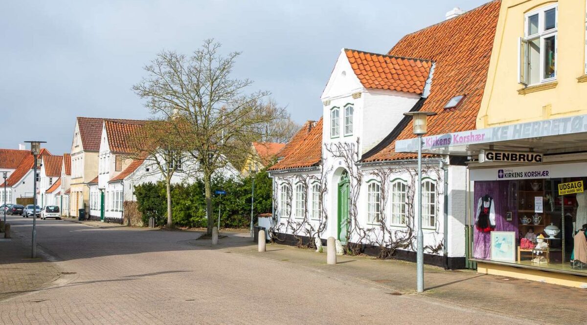 Foto af en gade med et gult hus med en forretning til højre. Ved siden af ses et lille hvidt hus med planter kravlende op af facaden. Taget er rødt ligesom de andre bygningers i gaden