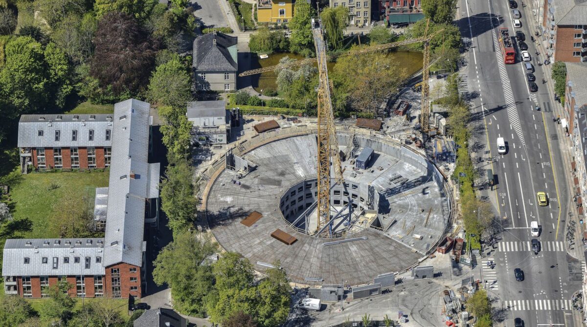 Dronefoto af en cirkelrund byggeplads dækket af et beton"låg". Midt på pladsen står en høj, gul kran.